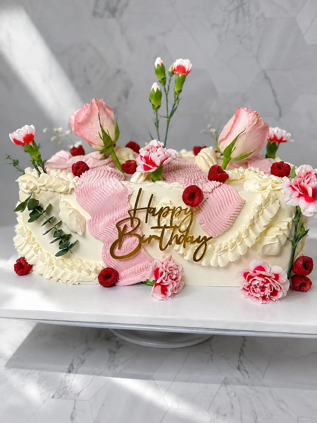 Torta floral con crema en tonos rosados, flores frescas y topper dorado de cumpleaños.